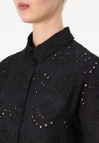 Camicia nera ricamata con collo a punta, caratterizzata da motivi decorativi a taglio e bottoni sul davanti. Texture morbida.