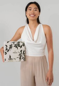 Weißes ärmelloses Oberteil mit drapiertem Ausschnitt; Model hält eine quadratische Box mit floralen Schwarz-Weiß-Muster und dem Schriftzug "PURELEI" darauf. Neutrale Hose.