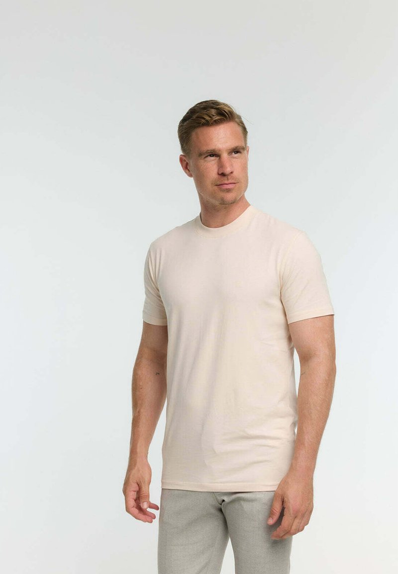 Camiseta beige de manga corta hecha de tela suave, con cuello redondo y un diseño ajustado, combinada con pantalones gris claro.