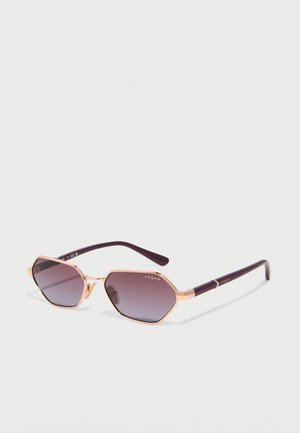 Hexagonal sunglasses with rose gold metal frame and dark purple gradient lenses, purple arms, and "Vogue" logo on lenses and arms.