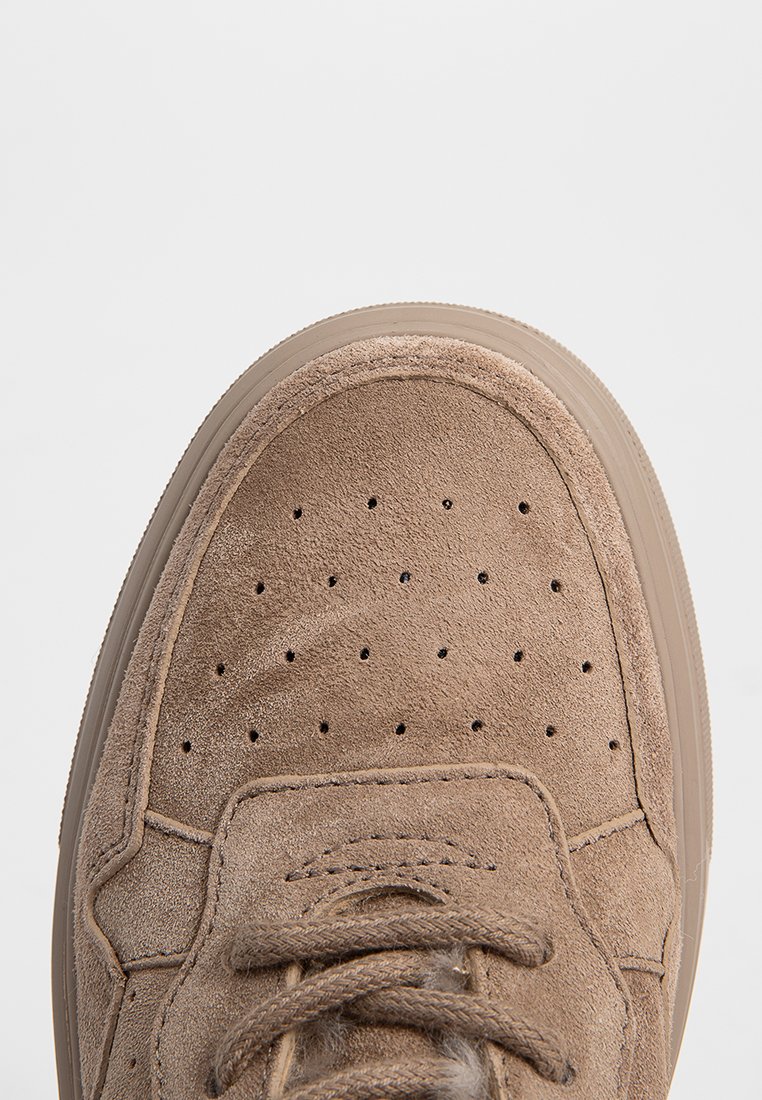 Braune Wildleder-Sneaker mit durchbrochenen Details, flachen Schnürsenkeln und einer strukturierten Gummisohle. Das Design ist low-cut mit minimalem Branding.