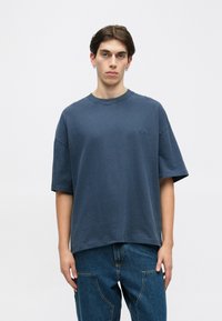 Mladý muž s krátkými tmavými vlasy má na sobě oversized modré tričko a volné modré džíny, stojí na pozadí s jednoduchým světlým pozadím.