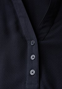 Chemise bleu marine foncé avec un décolleté en V et un tissu texturé. L'avant présente trois boutons noirs avec des motifs circulaires.