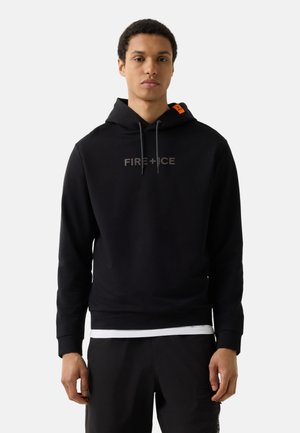 Jonge man die een zwarte hoodie draagt met de tekst "FIRE + ICE" en een oranje label op de capuchon, zwarte broek en een wit ondershirt.