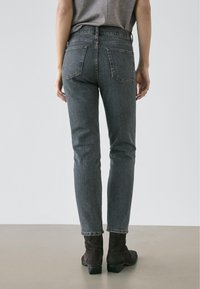Personne portant un jean en denim gris taille haute et des bottines noires, debout sur un sol beige devant un fond blanc uni.