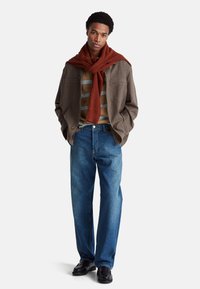 Hnědá vlněná bunda přes béžové pruhované tričko, džíny a černé boty. Červený svetr je přehozený přes krk.