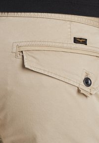 Beige cargo pants with a side pocket, featuring a black button and a stitched brand label. Smooth fabric texture and tailored cut.