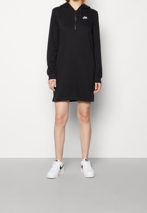 Žena oblečená v černých dlouhých rukávech s kapucí Nike ve formě šatů se zipem vpředu a bílé tenisky Nike s černými logy Swoosh.