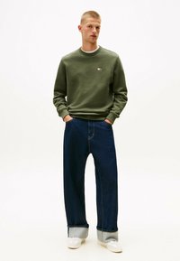 Felpa verde oliva con collo rotondo e piccolo logo, abbinata a jeans blu scuro larghi con risvolti e sneakers bianche.