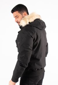 Veste noire avec capuche bordée de fourrure, dotée d'une fermeture éclair, de poignets côtelés et de poches latérales. Tissu lisse avec détails de couture visibles.