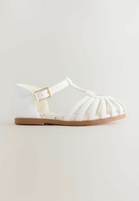White sandal made of smooth synthetic material featuring T-strap design, cut-out details, and a gold buckle. Light brown rubber sole.