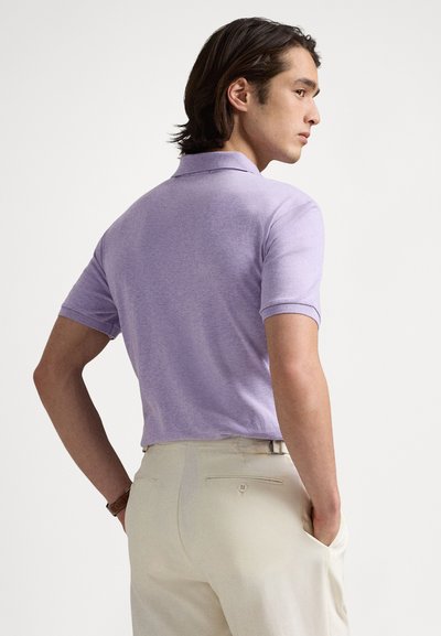 Ανοιχτό μωβ πόλο πουκάμισο με κοντά μανίκια, κατασκευασμένο από αεριζόμενο ύφασμα, συνδυασμένο με παντελόνι σε κρεμ απόχρωση και διακριτική λεπτομέρεια πίσω τσέπης.