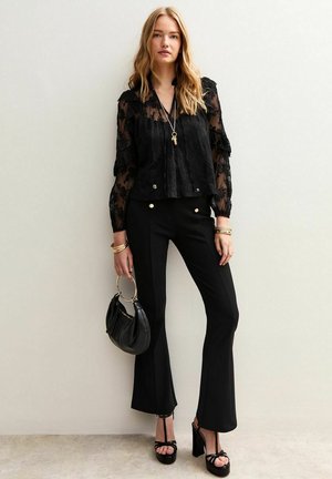 Zwarte kanten blouse met ruches aan de mouwen, gecombineerd met hoge zwarte wijde broeken met gouden knopen. Accessoires omvatten een zwarte tas en sieraden.