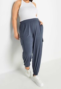 Kvinna iklädd en vit ärmlös topp och lösa mörkblå cargojoggingbyxor med vita sneakers, stående mot en vit bakgrund.