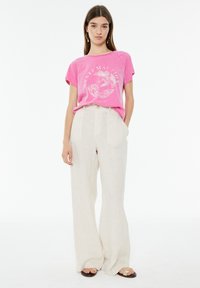 Camiseta de algodón rosa de manga corta con un diseño gráfico, combinada con pantalones de lino color crema de pierna ancha. Sandalias con suela plana.