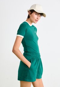 Ung person iført en grøn kortærmet shirt med hvid kant, matchende grøn shorts med elastisk talje og en beige baseball cap, hænderne i lommerne.