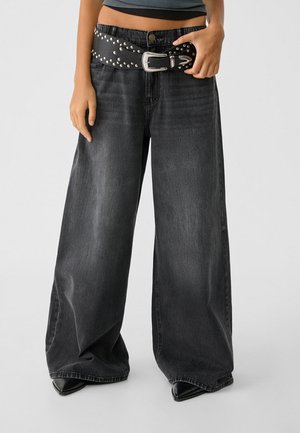 Jean noir délavé à jambes larges porté avec des chaussures noires pointues, une ceinture noire cloutée et un débardeur court sombre, montré de la moitié du torse vers le bas.