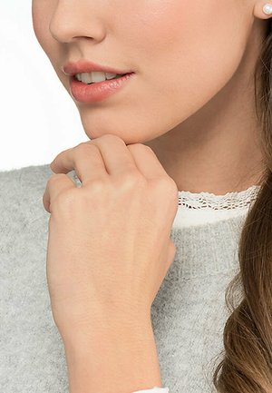 Nahaufnahme einer Hand, die auf einem grauen Pullover ruht, mit einem sichtbaren weißen Spitzenkragen. Der Hautton wirkt hell. Die Haare sind braun und locker.