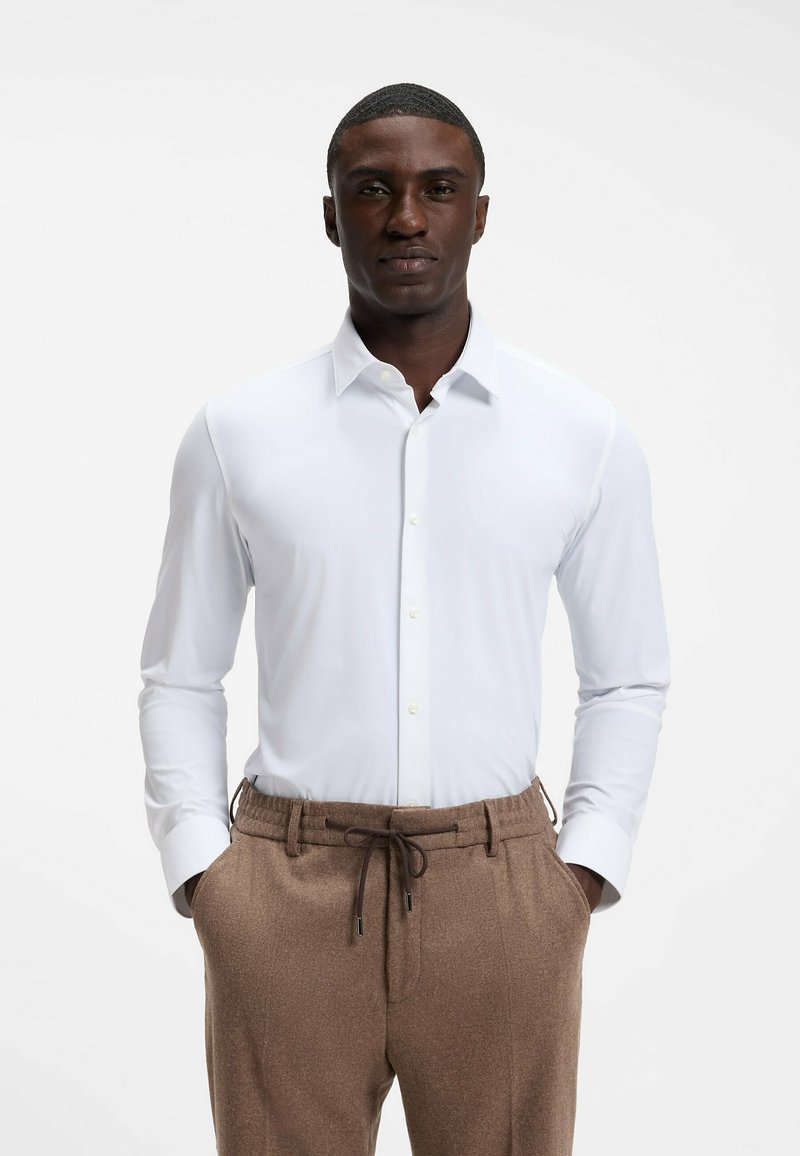 Homme debout portant une chemise blanche cintrée à boutons et un pantalon marron à cordon, les mains dans les poches, devant un fond clair uni.