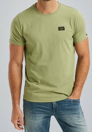 Homme portant un t-shirt PME Legend à manches courtes vert clair avec un écusson logo noir, associé à un jean bleu et une main dans la poche.