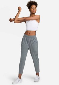 Lichtgrijze sportbroek met elastische tailleband, gecombineerd met een wit crop topje. Model dat stretcht, draagt witte sneakers; gladde stoftextuur.