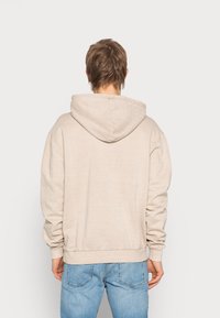 Beige hettegenser med avslappet passform, ribbestrikkede mansjetter og kant, samt glatt tekstur. Brukt sammen med lyseblå jeans.