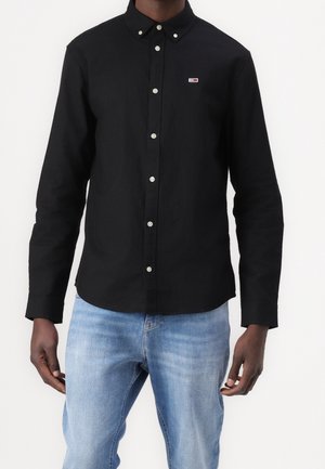 Homme portant une chemise noire boutonnée avec un petit logo rouge et blanc sur la poitrine, associée à un jean bleu clair sur un fond uni.
