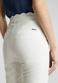 Personne portant un pantalon blanc taille haute avec une ceinture festonnée et une petite étiquette métallique de la marque sur la poche arrière, associé à un top bleu foncé.