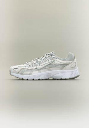 Zapatilla de running Nike blanca y gris claro con malla y superposiciones sintéticas, diseño con cordones y suela gruesa texturizada sobre fondo liso.