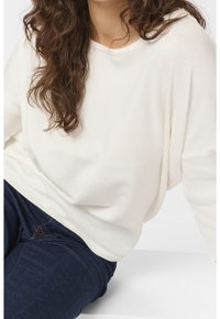 Personne aux cheveux bouclés portant un pull blanc ample et un jean bleu foncé, assise avec un bras reposant sur une surface blanche.