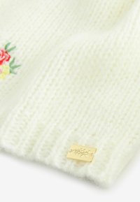 Valge kootud kangas, millel on väikesed tikitud roosad ja kollased lilled ning kuldne ristküliku kujuline silt lillegraveeringuga.