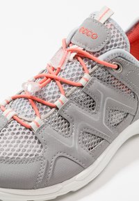 Scarpa atletica leggera grigia con tessuto in mesh, lacci elasticizzati corallo a contrasto e linguetta imbottita. Presenta dettagli stampati e una suola bianca.