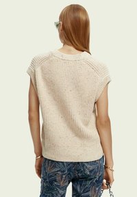 Haut beige en tricot à manches courtes avec un motif texturé. Porté avec un pantalon bleu à motifs, offrant une coupe décontractée.