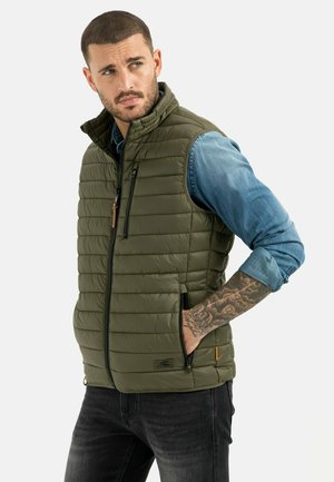 Olivgrüne gesteppte Weste mit Stehkragen, durchgehendem Reißverschluss und seitlichen Taschen. Getragen über einem Jeanshemd, zeigt ein sichtbares Tattoo am Arm.
