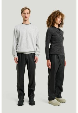 Mand i grå sweatshirt og sorte bukser står ved siden af kvinde i mørkegrå knappet bluse og sorte bukser på lys baggrund.