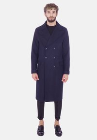 Cappotto peacoat in lana navy con design doppiopetto, ampie alette, grandi bottoni e tasche frontali; abbinato a pantaloni neri aderenti e scarpe slip-on.