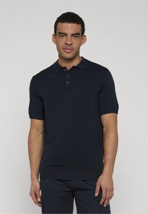 Junger Mann mit kurzen, lockigen Haaren, der ein dunkelblaues Poloshirt mit kurzen Ärmeln und dunkle Hosen trägt, vor einem schlichten Hintergrund stehend.