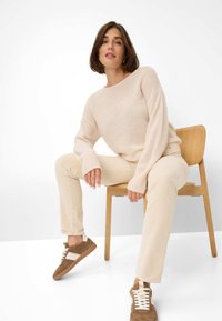Beige Wollpullover, cremefarbene Hose und braune Sneakers mit weißen Akzenten. Sitzend auf einem Holzstuhl vor einem schlichten Hintergrund.