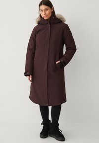 Lange bordeauxrode parka met een capuchon gevoerd met bont, ritssluiting met drukknopen, zijzakken en zwarte accenten. Draagt deze over zwarte legging en laarzen.