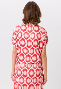 Blouse à motifs géométriques rouge et blanc avec des manches courtes, présentant une texture douce et lisse ainsi qu'une coupe décontractée.