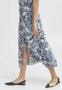 Robe maxi bleu clair avec impression paisley marine, dotée d'une fente sur le côté et d'une coupe décontractée. Portée avec des ballerines pointues noires et beiges.