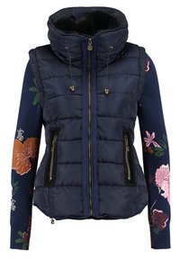 Veste matelassée bleu marine avec col montant et fermetures éclair frontales sur un pull à manches longues fleuri avec des motifs floraux multicolores sur les manches.