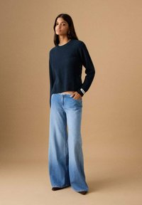 Femeie în picioare cu mâna în buzunar purtând un pulover bleumarin închis și blugi largi albastru deschis, pe un fundal bej.