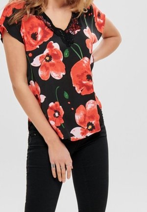 Femme portant un chemisier noir à large imprimé floral rouge et rose et un pantalon noir, une main sur la hanche et l’autre pendant.