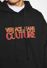 Svart huvtröja med snörning; har djärv, flerfärgad broderi som visar "VERSACE JEANS COUTURE" i gult och rött. Mjuk textur.
