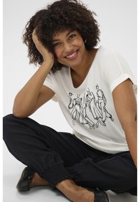 T-shirt en coton blanc avec un design en dessin noir, associé à un pantalon noir décontracté. Le modèle est assis avec une main sur sa joue.