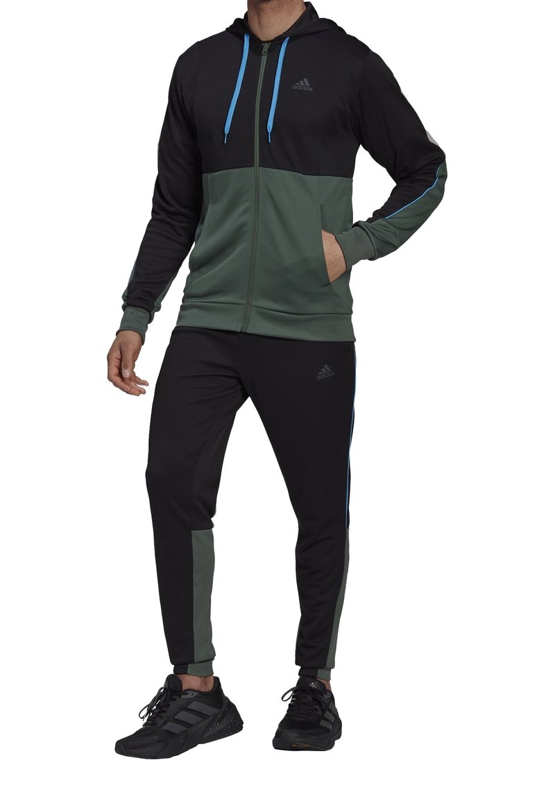 Homme portant une veste à capuche Adidas noire et verte et un pantalon Adidas noir et vert assorti avec des baskets Adidas noires.