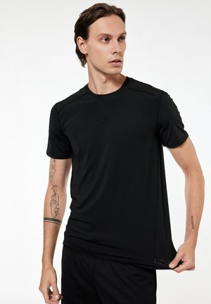 Jonge man met halflang haar die een effen zwart t-shirt met korte mouwen draagt, trekt de zoom van het shirt weg van zijn lichaam, waardoor tatoeages op beide armen zichtbaar zijn.