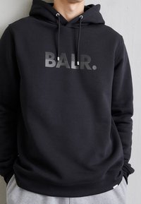 Sudadera negra de tela suave; cuenta con el logo gris "BALR.", cordones ajustables y un bolsillo tipo canguro en la parte frontal.