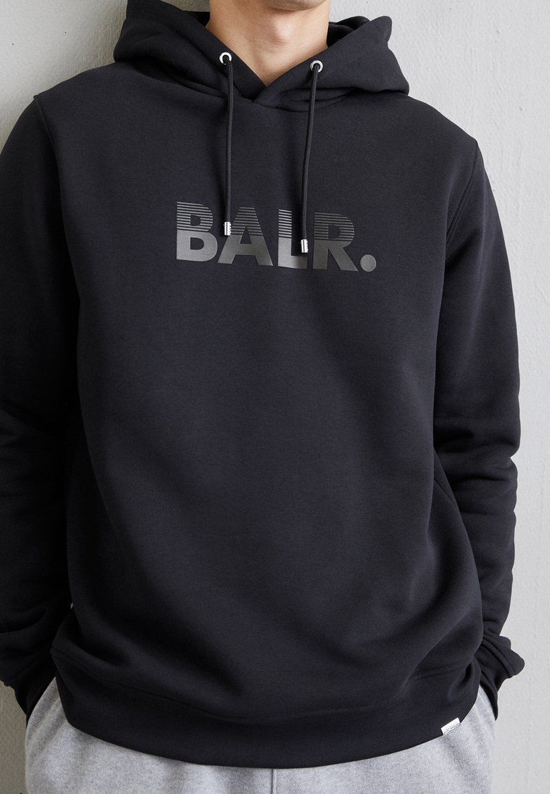 Sudadera negra de tela suave; cuenta con el logo gris "BALR.", cordones ajustables y un bolsillo tipo canguro en la parte frontal.